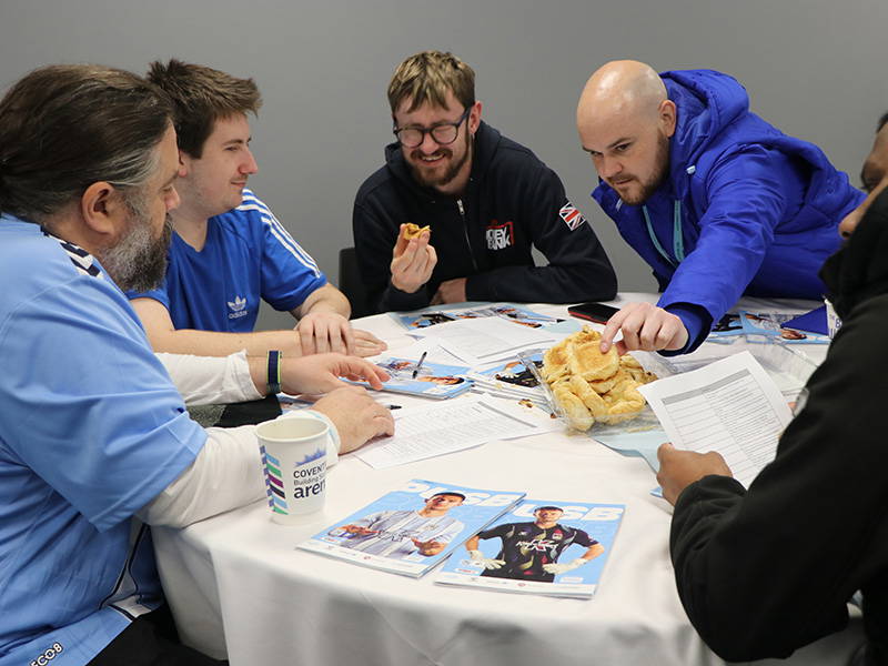 This image shows four men sat around a table, the first man has brown hair and is wearing a short sleeved blue t-shirt with a white long sleeved t-shirt underneath, they are sat nearest the camera. Next to this man is another man who is also brunette and is wearing a short sleeved blue t-shirt. The third person in this photo is a man with brown hair, black rectangular glasses and is wearing a black coat. Finally, there is the coach George who is wearing the CCFC royal blue coat, he is reaching for the snacks in the middle of the table. There are programmes with Brad Collins and Bobby Thomas on the front covers.